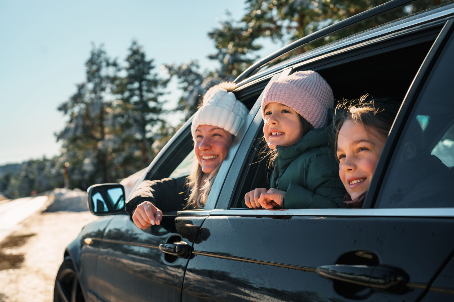 Three children in winter hats leaning out of car windows, smiling, with evergreen trees behind them