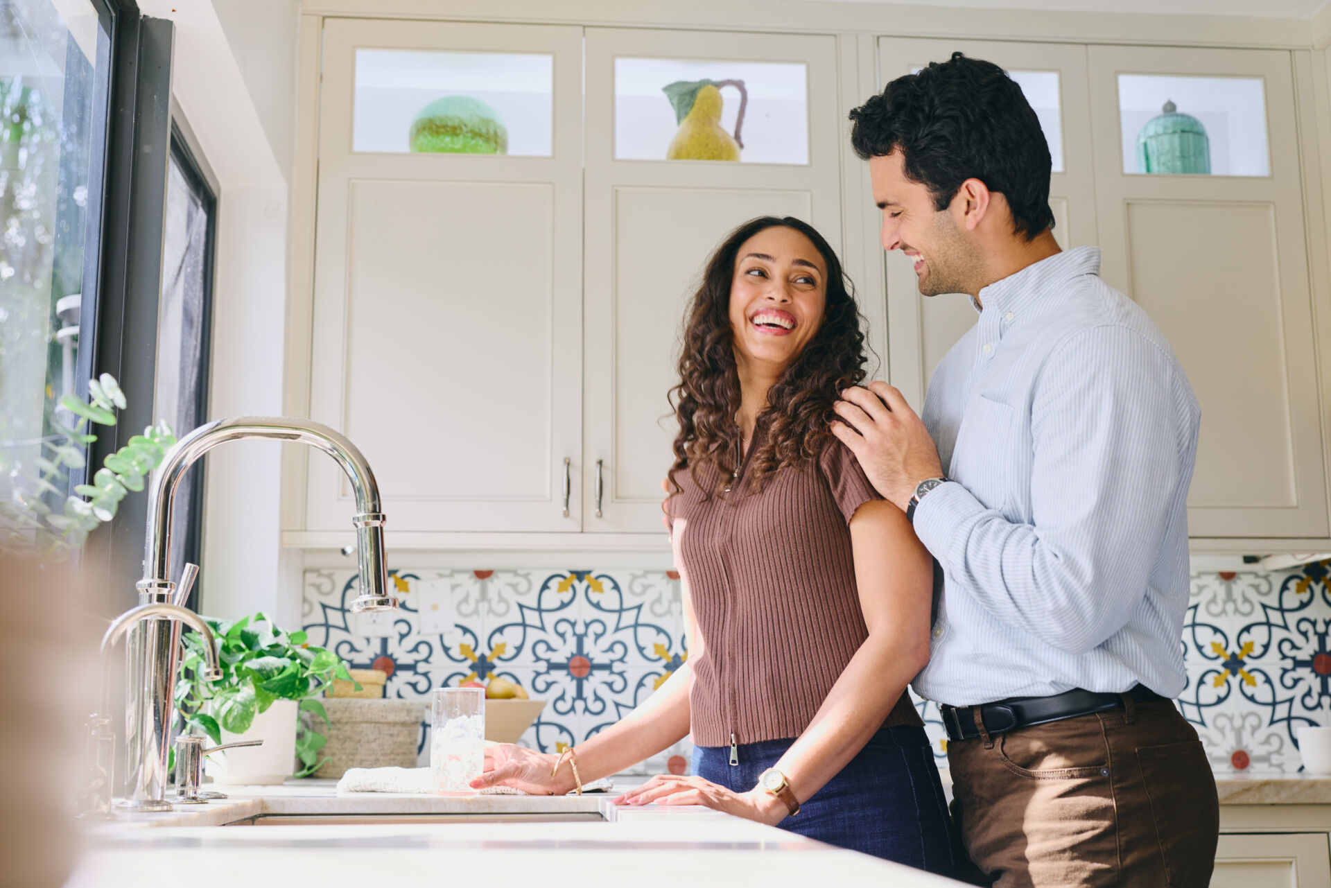 Couple smiling at each other in a bright kitchen with patterned tile backsplash