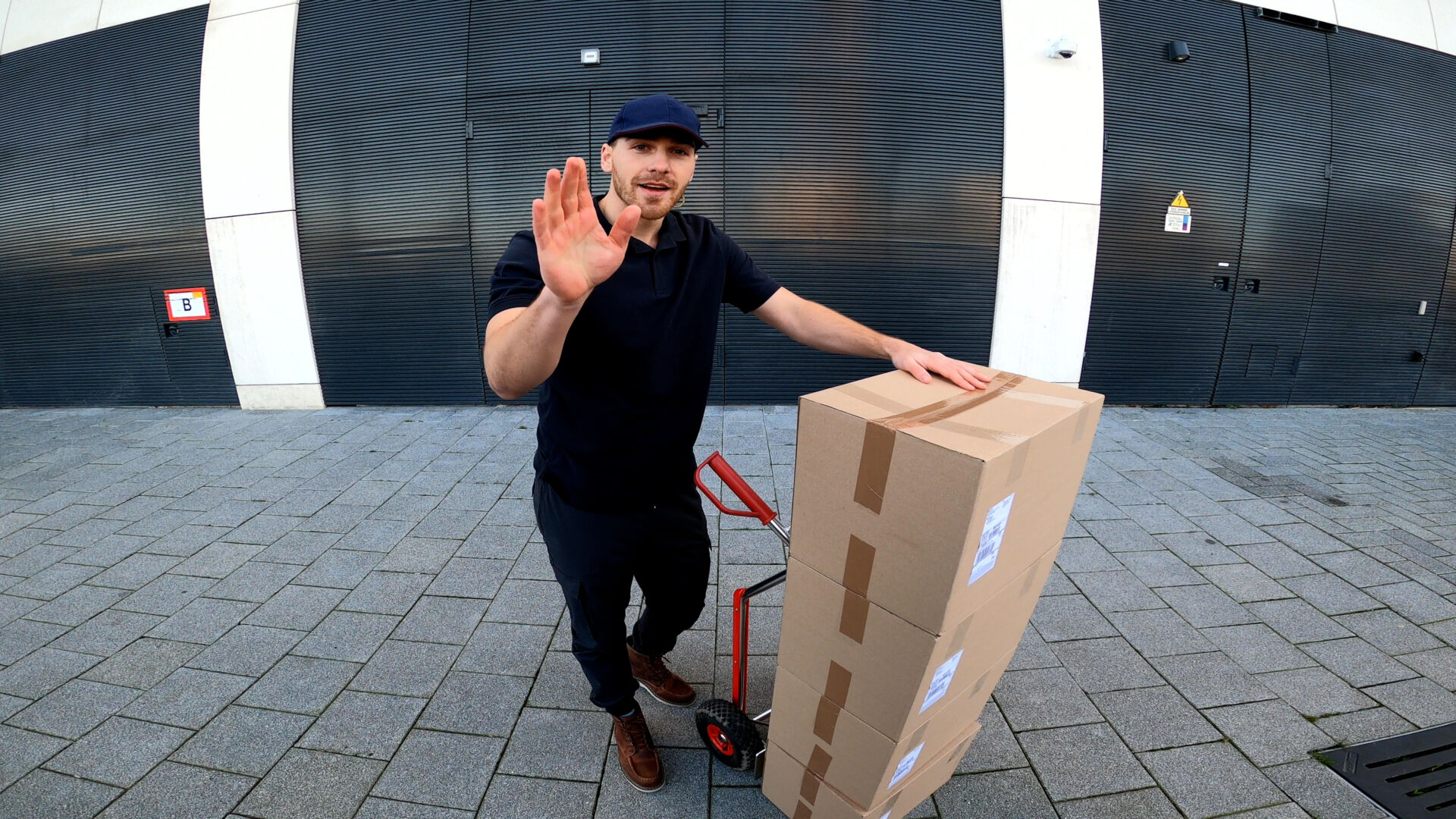 Delivery worker with a hand truck of boxes waving outside a store entrance