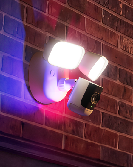 Security camera with an activated floodlight and blue and red warning lights mounted on a brick building exterior