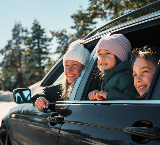 Three children in winter hats leaning out of car windows, smiling, with evergreen trees behind them