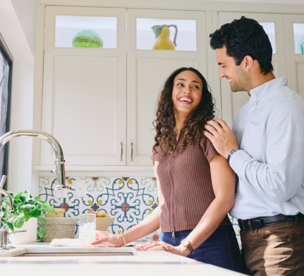 Couple smiling at each other in a bright kitchen with patterned tile backsplash