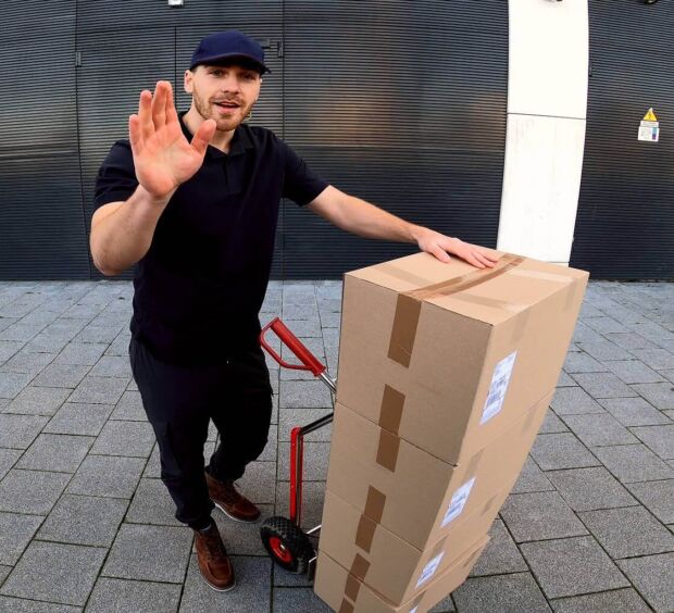 Delivery worker with a hand truck of boxes waving at a camera outside a back entrance